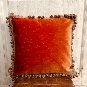 Könnte beinhalten: Ein orangefarbenes Samtkissen mit dekorativem Quastenbesatz. Das Kissen liegt auf einer Holzoberfläche.