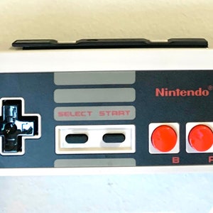 May include: A black and white Nintendo NES controller with red buttons. The controller has a gray directional pad, two gray buttons labeled "Select" and "Start", and two red buttons labeled "B" and "A".
