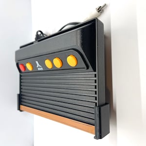 May include: A black Atari 2600 video game console with a woodgrain trim. The console has a row of five yellow buttons and a red button. The console is mounted on a wall.