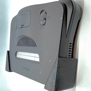 May include: A gray Nintendo 64 console with a black plastic base and a slot for game cartridges. The console has a vent on the top and a power button on the front.