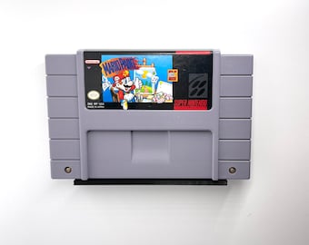 Puede incluir: Un cartucho de juego Super Nintendo Entertainment System para Mario Paint. El cartucho es gris con una etiqueta negra que presenta una ilustración colorida de Mario pintando. El texto "Mario Paint" está impreso en blanco en la etiqueta.