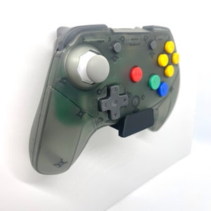 Puede incluir: Un mando de Nintendo 64 translúcido gris con un soporte de pared negro. El mando tiene una palanca analógica gris, un pad direccional negro y botones de colores.