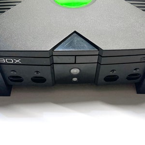 Puede incluir: Una consola de videojuegos Xbox negra y gris. La consola tiene un bot&oacute;n de encendido verde y la palabra "BOX" es visible. La consola est&aacute; sobre una superficie blanca.