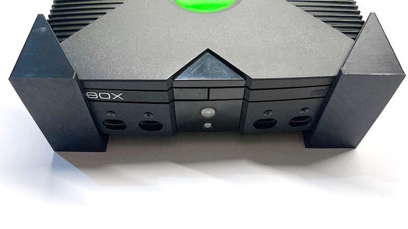Puede incluir: Una consola de videojuegos Xbox negra y gris. La consola tiene un bot&oacute;n de encendido verde y la palabra "BOX" es visible. La consola est&aacute; sobre una superficie blanca.