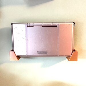 May include: A pink Nintendo DS handheld gaming console with a silver faceplate and the Nintendo logo. The console is in a brown plastic stand.