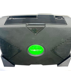 Puede incluir: Consola Xbox negra con un bot&oacute;n de encendido circular verde y el logotipo de Xbox. La consola tiene forma rectangular con una rejilla de ventilaci&oacute;n en la parte superior y un puerto USB. Un dispositivo de juego cl&aacute;sico.