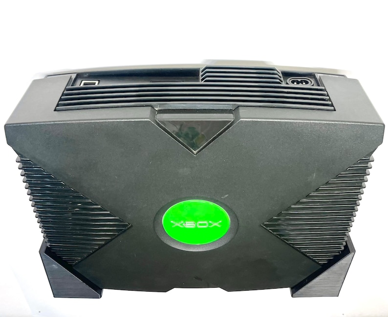 Puede incluir: Consola Xbox negra con un bot&oacute;n de encendido circular verde y el logotipo de Xbox. La consola tiene forma rectangular con una rejilla de ventilaci&oacute;n en la parte superior y un puerto USB. Un dispositivo de juego cl&aacute;sico.
