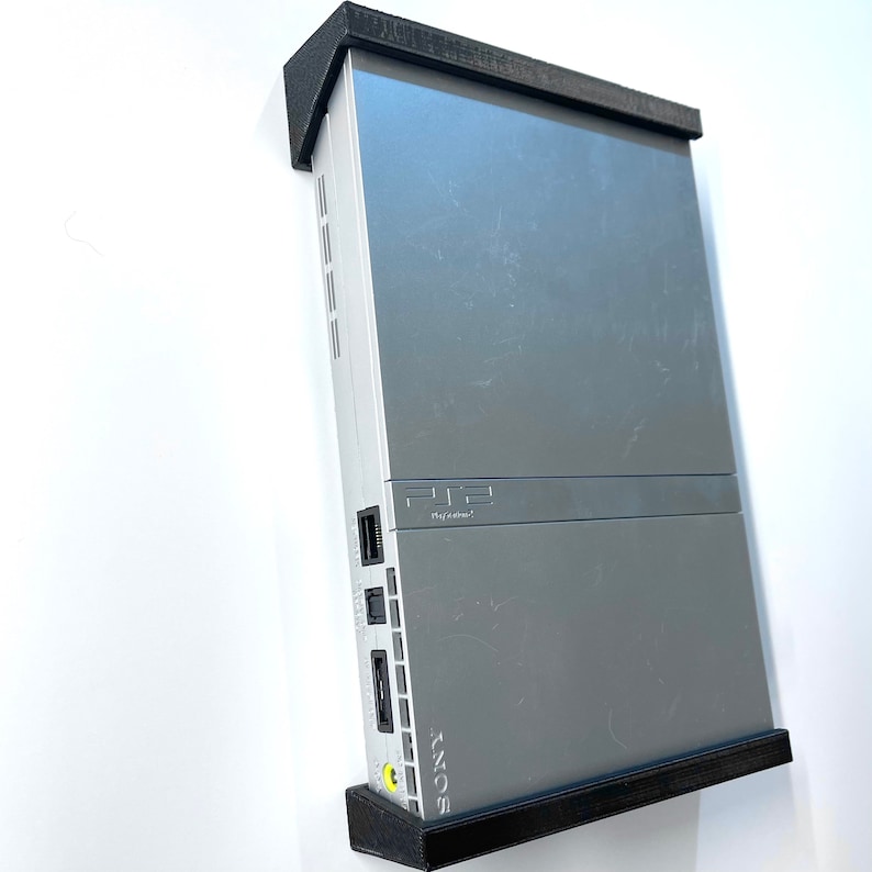 K&ouml;nnte beinhalten: Eine silberne PlayStation 2 Konsole, die an einer schwarzen Wandhalterung montiert ist. Die Konsole hat eine schwarze Frontplatte mit dem PlayStation 2 Logo und dem Text "Netzwerkadapter" darauf gedruckt. Die Konsole ist auch mit "SONY" in wei&szlig;en Buchstaben beschriftet.