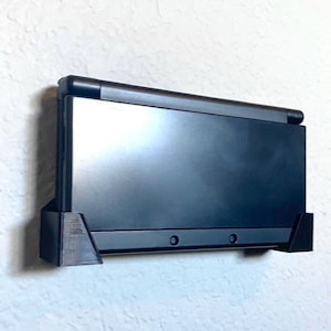 May include: A black wall-mounted console, likely for a gaming device, is displayed against a textured white wall. The console is rectangular with a sleek, modern design, featuring a flat screen and a supportive base.