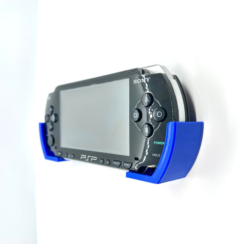 Peut inclure: Une console de jeu portable Sony PlayStation Portable (PSP) noire avec un support mural bleu. La PSP est mont&eacute;e au mur avec l'&eacute;cran tourn&eacute; vers l'avant. La PSP a un bouton d'alimentation bleu et un bouton de maintien bleu.