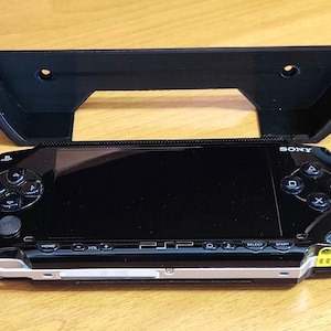 Peut inclure: Une console de jeu portable Sony PlayStation Portable (PSP) noire avec un support mural en plastique noir. La PSP a un &eacute;cran et des boutons noirs, et le support mural a deux trous pour le montage.