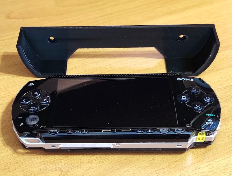 Peut inclure: Une console de jeu portable Sony PlayStation Portable (PSP) noire avec un support mural en plastique noir. La PSP a un &eacute;cran et des boutons noirs, et le support mural a deux trous pour le montage.