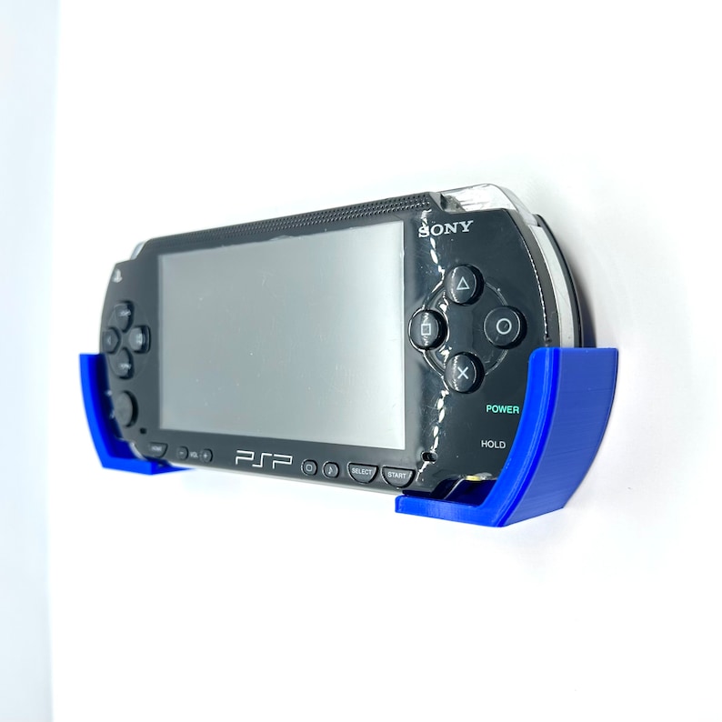 Peut inclure: Une console de jeu portable Sony PSP noire avec un support mural bleu. La console a un &eacute;cran, des boutons et un bouton d'alimentation intitul&eacute; "POWER".