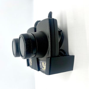 May include: A black wall mount for two Atari 2600 game controllers. The mount has a black plastic base and two black plastic arms that hold the controllers. The controllers have black knobs and a black cord.