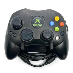以下が含まれることがあります： 緑色のXboxロゴとカラーボタンが付いた黒いXboxコントローラー。 コントローラーには方向パッド、2つのアナログスティック、いくつかのボタンがあります。 黒いケーブルがコントローラーに接続されています。
