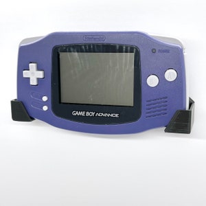 May include: A purple Game Boy Advance handheld console with a black screen, white buttons, and the text "GAME BOY ADVANCE" printed on the bottom. The console is held in a black 3D-printed stand. The Nintendo logo is visible.