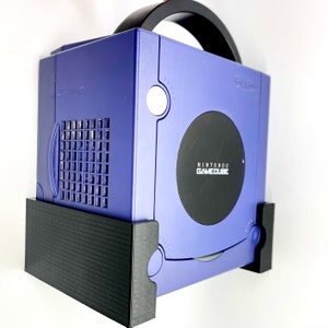 May include: A purple Nintendo GameCube console with a black handle and stand. The console has the words "NINTENDO GAMECUBE" printed on the front. The stand is black and holds the console securely.