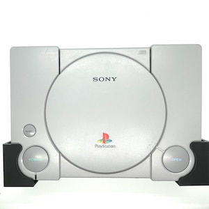 May include: Silver PlayStation console with the Sony logo and PlayStation logo. Features a disc tray, power and reset buttons, and an open button. The console is set on a black stand.