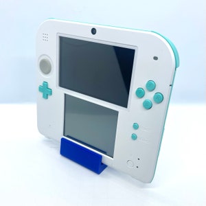 Puede incluir: Una consola de juegos portátil Nintendo 2DS blanca y azul claro con un soporte azul. La consola tiene una pantalla negra y una variedad de botones, incluidos A, B, X, Y, Start y Select.