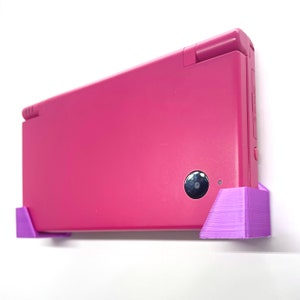 Puede incluir: Una consola de juegos portátil Nintendo DS Lite rosa con un soporte de pared impreso en 3D morado.