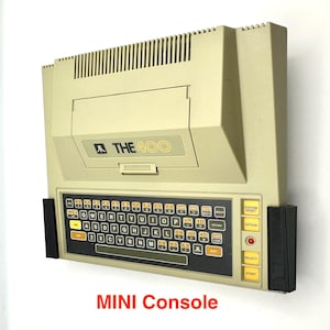 Può includere: Un computer Atari 400 beige con una tastiera nera e uno schermo bianco e nero. Il computer è montato su una parete con staffe nere. Il testo "MINI Console" è stampato sotto il computer.