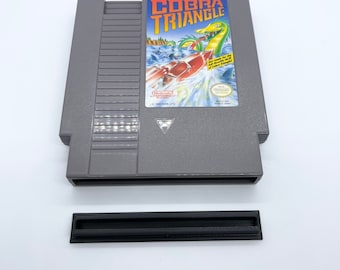 May include: A gray Nintendo Entertainment System (NES) game cartridge for Cobra Triangle. The label features a colorful illustration of a boat speeding through water with a large snake in the background. The text "Cobra Triangle" is printed in white letters on a black background.