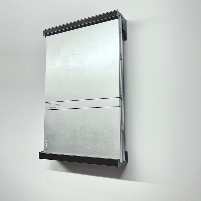 K&ouml;nnte beinhalten: Eine silberne PlayStation 2 Konsole mit einer schwarzen Frontplatte und einem schwarzen Rahmen. Die Konsole ist mit einer schwarzen Halterung an der Wand befestigt.