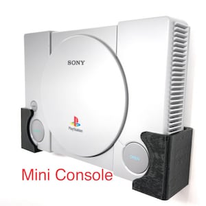 Puede incluir: Una consola Sony PlayStation blanca con un soporte de pared negro. La consola tiene una unidad de disco plateada y el texto "PlayStation" y "SONY" impreso en la parte delantera. El soporte de pared está hecho de plástico negro y tiene la palabra "OPEN" impresa en él.