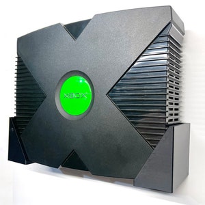 Könnte beinhalten: Schwarze Xbox-Spielekonsole mit grünem Power-Button und Xbox-Logo. Die Konsole hat ein markantes X-förmiges Design und eine gerippte Seitenwand.