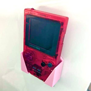 Può includere: Un supporto a parete rosa per una console per videogiochi portatile Game Boy Color rossa. Il supporto è in plastica e ha una forma triangolare. La Game Boy Color è nel supporto e guarda la fotocamera. La Game Boy Color ha uno schermo nero e una disposizione dei pulsanti rosso e nero.