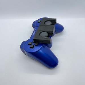 May include: A blue Sony PlayStation 2 controller with a black 3D printed attachment on top. The attachment has two circular openings.