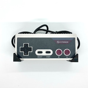 May include: A white and gray Hyperkin brand NES controller with a black directional pad, two gray buttons, and two red buttons labeled "A" and "B".
