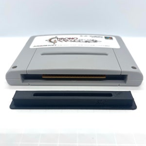 May include: A gray Super Nintendo Entertainment System (SNES) game cartridge with a brown label. The label has the text "CHRONO TRIGGER" in red and white. The cartridge is sitting on a white surface. A black plastic cartridge case is also shown.