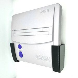 Peut inclure: Une console Super Nintendo Entertainment System blanche avec un support mural noir. La console a un logement de cartouche gris et deux boutons violets.