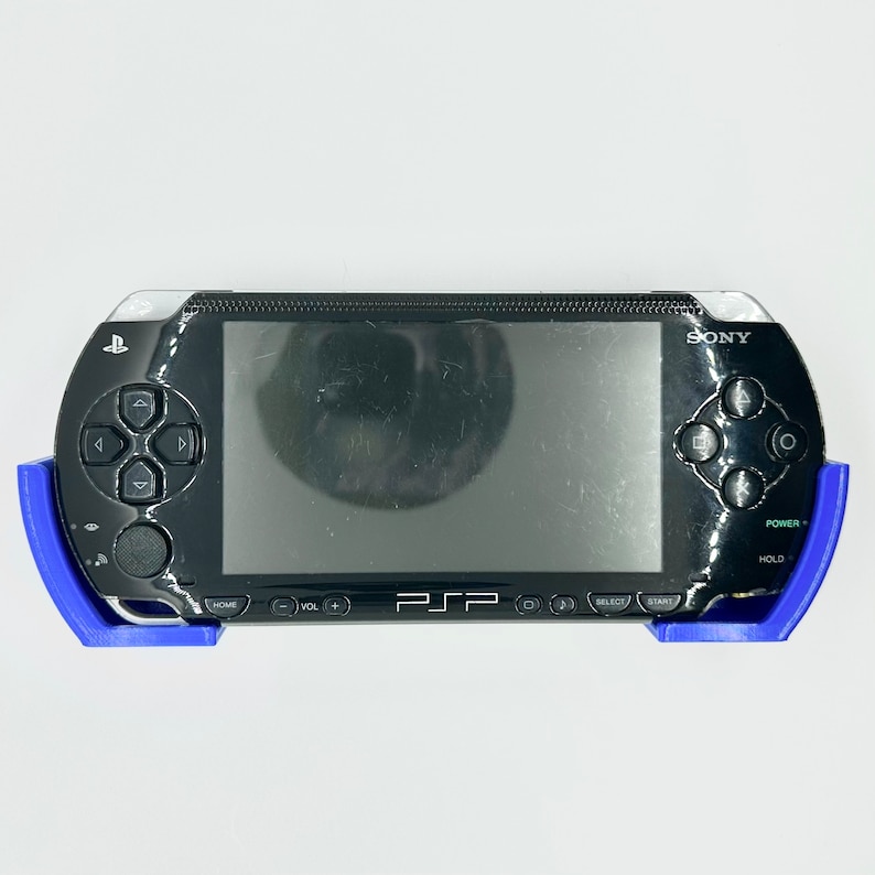 Peut inclure: Une console de jeu portable Sony PSP noire avec des coques de protection bleues sur les c&ocirc;t&eacute;s. La console a un &eacute;cran noir et des boutons, dont un bouton d'alimentation, un bouton de maintien, un bouton d'accueil, un bouton de s&eacute;lection, un bouton de d&eacute;marrage et des boutons de volume.