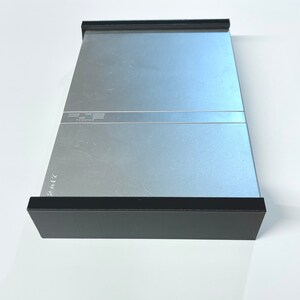 K&ouml;nnte beinhalten: Eine silberne PlayStation 2 Konsole mit schwarzem Rand. Die Konsole hat das PlayStation 2 Logo auf der Vorderseite.