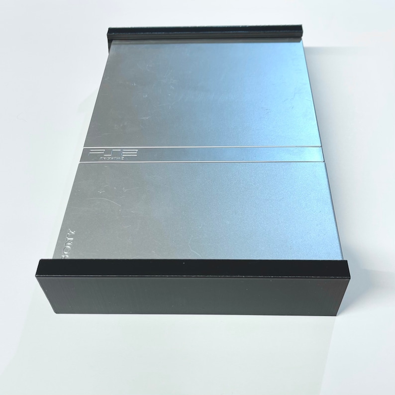 K&ouml;nnte beinhalten: Eine silberne PlayStation 2 Konsole mit schwarzem Rand. Die Konsole hat das PlayStation 2 Logo auf der Vorderseite.