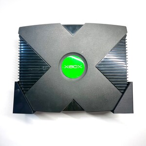 Puede incluir: Consola de videojuegos Xbox negra con un bot&oacute;n de encendido circular verde. La consola tiene un dise&ntilde;o distintivo en forma de X en la parte superior y un panel lateral acanalado. El logotipo de Xbox es visible en el bot&oacute;n de encendido.