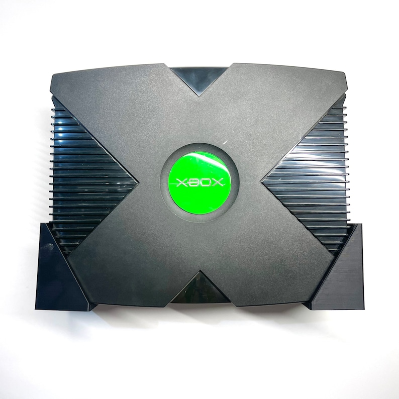 Puede incluir: Consola de videojuegos Xbox negra con un bot&oacute;n de encendido circular verde. La consola tiene un dise&ntilde;o distintivo en forma de X en la parte superior y un panel lateral acanalado. El logotipo de Xbox es visible en el bot&oacute;n de encendido.