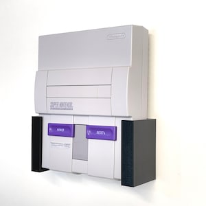 Puede incluir: Una consola Super Nintendo Entertainment System blanca con un soporte de pared negro y gris. La consola tiene un botón de encendido y reinicio morado.