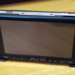 Peut inclure: Une console de jeu portable Sony PlayStation Portable (PSP) noire avec un protecteur d'&eacute;cran. La PSP est sur un support noir et poss&egrave;de une disposition de boutons noir et blanc.