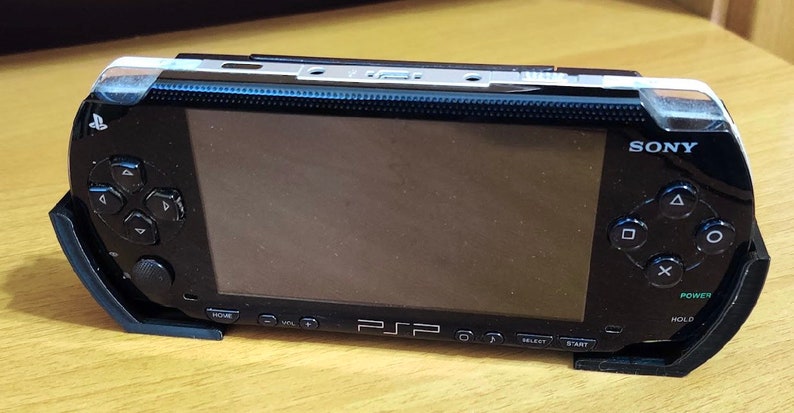 Peut inclure: Une console de jeu portable Sony PlayStation Portable (PSP) noire avec un protecteur d'&eacute;cran. La PSP est sur un support noir et poss&egrave;de une disposition de boutons noir et blanc.