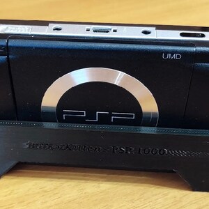 Peut inclure: Une console Sony PlayStation Portable (PSP) 1000 noire est plac&eacute;e dans un support noir imprim&eacute; en 3D. La PSP a un lecteur UMD argent&eacute; et un bouton circulaire argent&eacute; avec le logo PSP.