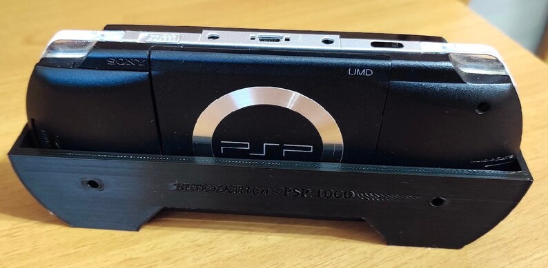 Peut inclure: Une console Sony PlayStation Portable (PSP) 1000 noire est plac&eacute;e dans un support noir imprim&eacute; en 3D. La PSP a un lecteur UMD argent&eacute; et un bouton circulaire argent&eacute; avec le logo PSP.