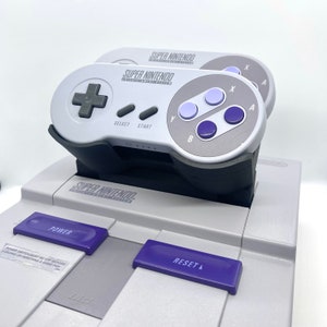 Op de afbeelding: Een grijze Super Nintendo Entertainment System-console met twee grijze controllers die aan de voorkant zijn bevestigd. De console heeft een paarse aan/uit-knop, een paarse resetknop en een grijze uitwerpknop. De tekst "SUPER NINTENDO" staat op de console gedrukt.