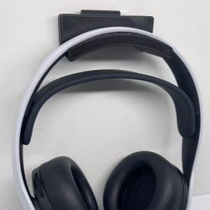 May include: A black headset with white accents is hanging on a black wall mount. The headset has a padded headband and large earcups.