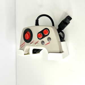 May include: A white NES Advantage joystick with red buttons and a black cord. The joystick has the words "NES Advantage" and "Nintendo" printed on it. The joystick is mounted on a white plastic wall mount.