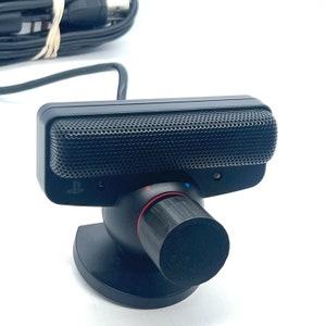 Puede incluir: Una cámara PlayStation Eye negra con un micrófono y un soporte. La cámara tiene un diseño negro y plateado con un logotipo de PlayStation.