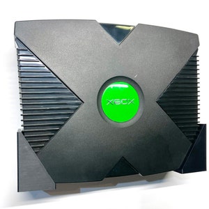 Puede incluir: Consola de videojuegos Xbox negra con un bot&oacute;n de encendido verde. La consola tiene un dise&ntilde;o distintivo en forma de X en la parte superior y un panel lateral acanalado. La palabra "XBOX" est&aacute; impresa en verde en el bot&oacute;n de encendido.
