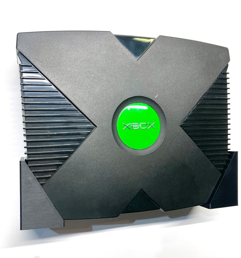 Puede incluir: Consola de videojuegos Xbox negra con un bot&oacute;n de encendido verde. La consola tiene un dise&ntilde;o distintivo en forma de X en la parte superior y un panel lateral acanalado. La palabra "XBOX" est&aacute; impresa en verde en el bot&oacute;n de encendido.